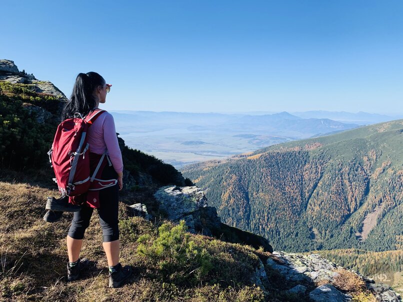 The person with the red backpack is standing on the top of the mountain, looking at the picturesque landscape with valleys and mountains in the distance.